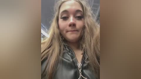 Media: Video of a young woman with long, messy blonde hair, wearing a black leather jacket, smiling slightly. Background is blurred, suggesting an indoor setting.