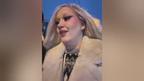 Media: Video of a blonde woman with long hair, wearing a white fur coat and black and white patterned scarf, smiling with a confident expression. Background shows a blurred blue sky and cityscape.