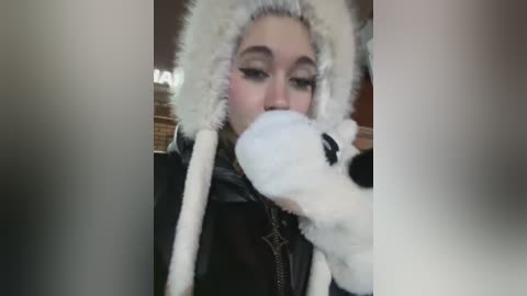 Media: Video of a young woman with light skin, wearing a white fur hood and black jacket, kissing a plush toy in a dimly lit room with brick walls.