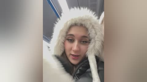 Media: Video of a young woman with fair skin and brown hair, wearing a white fur-trimmed hood and black jacket, looking up with a playful expression in a subway car.