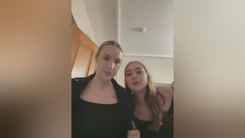 Media: A video shows two young women, one with light skin and blonde hair, the other with light skin and long brown hair, smiling closely together, wearing black tops. They stand in a modern, white-walled room.