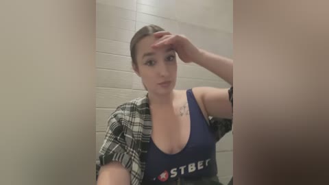 Media: Video of a fair-skinned woman with dark hair in a ponytail, wearing a blue sports bra and a plaid shirt, sitting in a public bathroom stall, touching her head.