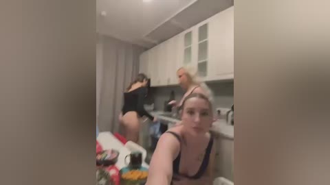 Media: A blurry, candid photo of three women in a modern kitchen, one bending over a counter, another facing the camera in a black bra, and the third partially visible.