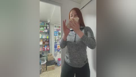 Media: Video of a woman with long red hair, wearing a grey hoodie and black pants, taking a selfie in a small, cluttered room with shelves of colorful products and scattered boxes.