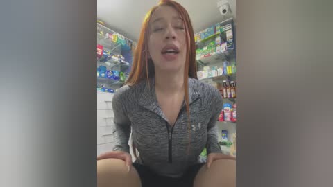 Media: Video of a young woman with long red hair and fair skin, wearing a grey zip-up hoodie, sitting on the floor in a brightly lit pharmacy, with shelves filled with various products in the background.