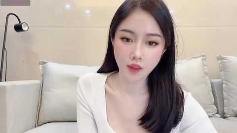 Media: Video of an East Asian woman with long black hair, fair skin, and a slender physique, wearing a white top, sitting on a beige couch with light-colored pillows. Background features a minimalist, modern room with a black lamp and cream-colored walls.