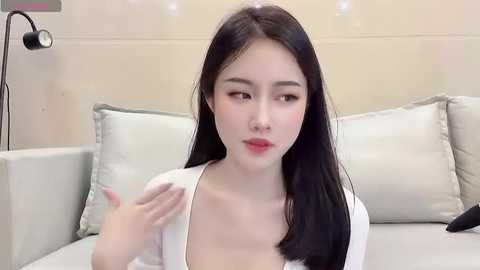 Media: Video of an East Asian woman with fair skin and long black hair, wearing a white top, sitting on a bed with white pillows in a modern, minimalist room.