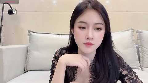 Media: Video of an East Asian woman with fair skin, long black hair, and delicate features, wearing a black lace top, sitting on a white couch with beige pillows. The background is a beige wall with a black floor lamp.