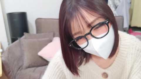 A video of an East Asian woman with short brown hair, wearing glasses, a white mask, and a cream-colored knitted sweater, sitting on a beige sofa with pink cushions, blurred background.