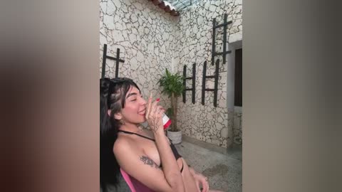 Media: A video of a smiling, topless Latina woman with long black hair, medium breasts, and a tattoo, sitting on a pink chair, drinking from a red cup, against a stone wall with black metal decor.