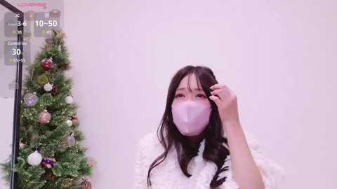 Video of an East Asian woman with long black hair, wearing a white fluffy sweater and a pink face mask, standing in front of a decorated Christmas tree, with a calendar showing 10:50 in the background.