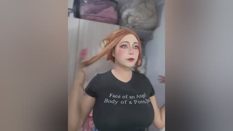 Media: Video of a fair-skinned woman with auburn hair, wearing a black t-shirt with the phrase \"Face of an Angel Body of a Pornstar\" in white letters. Background shows a messy closet with various clothes and bags.