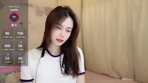 Media: Video of a young Asian woman with straight, long black hair, fair skin, and a petite frame, wearing a white T-shirt. She stands in a room with beige curtains and a pink toy on the left.