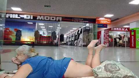 Media: Video of an elderly woman with short gray hair, lying on a patterned cushion in a shopping mall, wearing a blue shirt and red pants. Background shows retail stores and a vibrant display window.