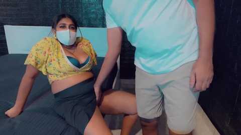 Media: Video of a Latina woman in a yellow floral top, blue bra, and gray skirt, wearing a mask, being held by a man in a white shirt and khaki shorts in a dimly-lit room with dark walls.