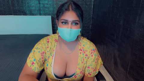 Media: Video of a curvy, light-skinned South Asian woman with large breasts, wearing a yellow floral dress with a plunging neckline, blue surgical mask, and sitting indoors with dark curtains and white walls.