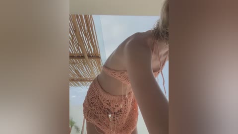 Media: Video of a blonde woman in a peach crochet bikini, leaning forward, with a straw roof in the background, sunlit and outdoors.