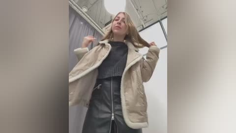 Media: Video of a young Caucasian woman with fair skin and blonde hair, wearing a beige fur-trimmed coat over a black sweater and leather skirt, adjusting her hair in a dimly lit, modern indoor space.