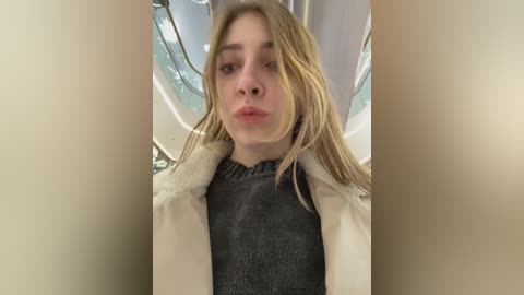 Media: Video of a young woman with long blonde hair, wearing a dark grey turtleneck and a white fur-trimmed coat. She has a neutral expression, standing in an indoor setting with modern, metallic railings in the background.