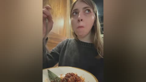 Media: A video of a young woman with light skin and long blonde hair, wearing a dark sweater, holding a bowl of stir-fried vegetables, seated indoors with wooden cabinets in the background.