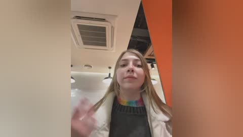 Media: A video of a young Caucasian woman with long blonde hair, wearing a colorful tie-dye shirt, standing in a modern, brightly lit room with orange and white walls, a white ceiling, and visible ventilation.