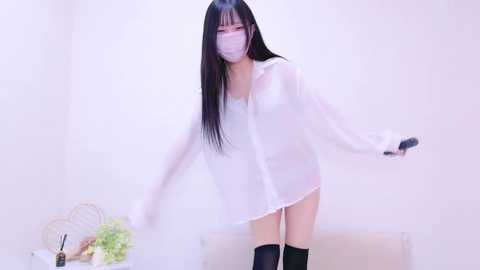 Media: Video of a slender, masked Asian woman with long black hair, wearing a sheer white blouse, black stockings, and holding a bouquet, standing against a plain white background.