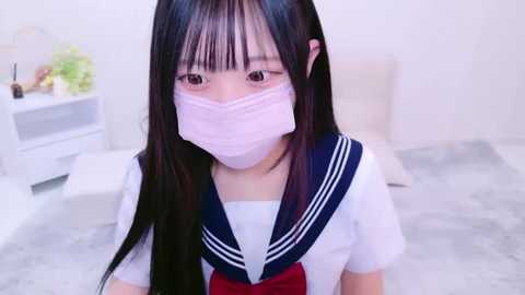 Media: A video of a young Japanese woman with long black hair, wearing a white schoolgirl uniform with a navy blue collar, pink face mask, and sitting on a light gray floor.