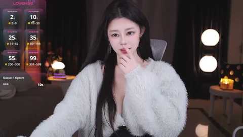 Media: Video of an East Asian woman with long black hair, wearing a white fur jacket, seated in a dimly lit room with a fireplace and candles.