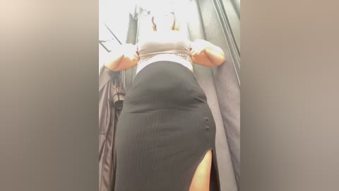 Media: Video of a woman with fair skin, medium build, and blonde hair, wearing a white crop top and a high-waisted black skirt with a high slit, standing in a dimly lit dressing room with gray walls and a mirror.