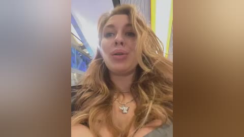 Media: Video of a young woman with long, wavy blonde hair and fair skin, wearing a black top. She has a medium build and is smiling, with a hint of a pout. Background shows a bright, modern interior with vertical blue and yellow stripes.