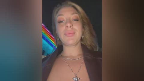 Media: Video of a fair-skinned woman with light brown hair, wearing a black jacket, smiling at the camera. She has a silver necklace with a pendant. Background includes a colorful rainbow light and blurred objects.