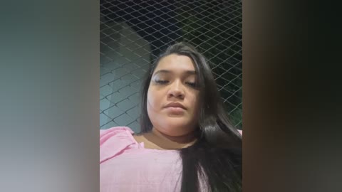 Media: Video of a young Latina woman with long, straight black hair, wearing a light pink blouse, standing in front of a chain-link fence in a dimly lit outdoor setting.