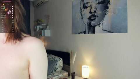 Media: Video of a woman's bare back, seen from behind, in a modern bedroom. She has light skin and shoulder-length brown hair. The room features a large, abstract painting of a woman's face, a bed with patterned bedding, and a lit white candle on a nightstand.