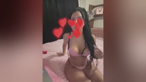 Media: Video of a woman with long black hair, wearing a pink bra and panties, kneeling on a pink bedspread. Red heart emojis cover her face. Bedroom setting with a wooden headboard, a dark curtain, and a white wall.