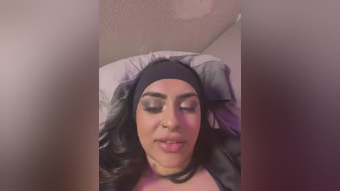 Media: A video of a young woman with medium skin tone, long dark hair, and a black headscarf, lying on a pillow in a dimly lit room. She has closed eyes and a serene expression.