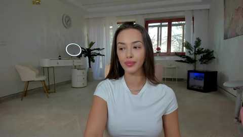 Media: Video of a young woman with straight, shoulder-length brown hair, wearing a white t-shirt, sitting in a modern, minimalist room with white walls, a round mirror, a desk, and potted plants.