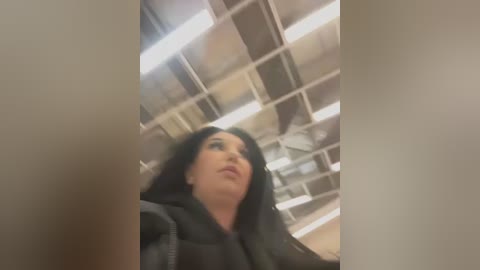 Media: Video of a young woman with long black hair, wearing a black jacket, standing in an office with fluorescent lighting and a ceiling fan.