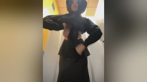 Media: Video of a woman in a black hooded sweatshirt and black pants, lifting the sweatshirt to reveal her small breasts. Background includes a yellow and blue wall, and white door.