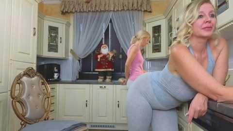 Media: Video of a blonde woman in a light blue dress bending over a kitchen counter, looking towards a blonde woman in a pink top and blue jeans. Kitchen with white cabinets and blue curtains.