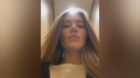 Media: Video of a young Caucasian woman with long, light brown hair, wearing a white top, taken from a low angle, highlighting her facial features. The background is blurred, suggesting an indoor setting with warm lighting.