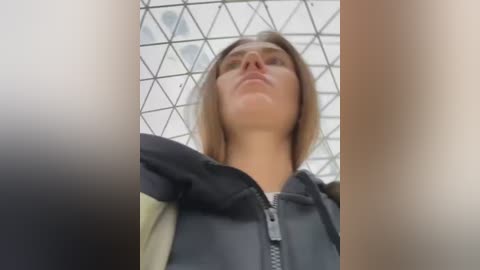 A low-angle video of a woman with shoulder-length brown hair, wearing a black and gray jacket, standing under a modern glass dome with a geometric pattern. The image has a slight blur on the sides.