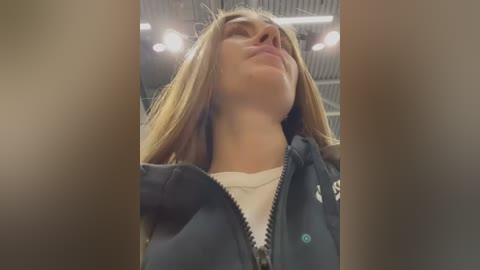 Media: A video of a young woman with straight, light brown hair, wearing a black jacket, standing in a gym with fluorescent lighting and metal beams in the background.