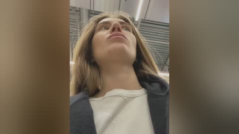 Video of a young woman with straight, shoulder-length brown hair, wearing a white ribbed sweater under a dark jacket, gazing upward with a contemplative expression in a modern, industrial room with metal beams and closed window blinds in the background.