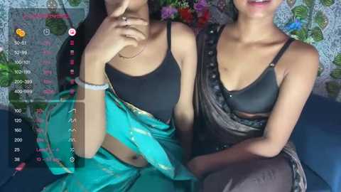 Media: Video of two South Asian women in black bras, one in a turquoise sari, seated against a colorful floral background.