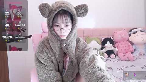 Media: Video of a young woman in a grey bear onesie, wearing glasses, sitting on a bed with plush toys. Background includes a screen showing a video feed and a pink blanket.