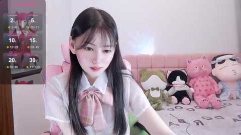 Media: Video of an Asian girl with long black hair, wearing a white shirt with a pink bow, sitting on a pink couch with plush toys.