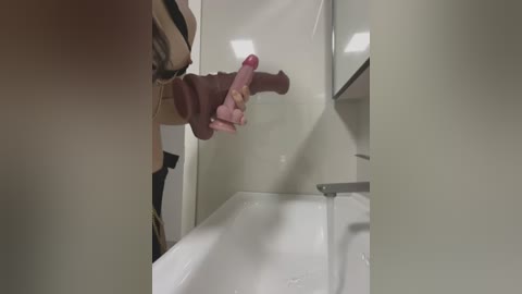 Media: Video of a person's hand holding a realistic, uncircumcised, brown dildo in a modern, white-tiled bathroom with a sink and mirror.