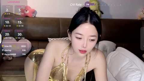 Media: A video of an East Asian woman with black hair in a ponytail, wearing a gold sequined dress, lying on a brown couch in a cozy room.