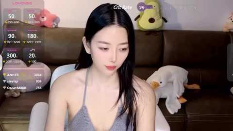 Media: Video of an East Asian woman with long black hair, wearing a gray halter top, sitting on a brown leather couch. Background includes stuffed toys, and digital game stats overlay.