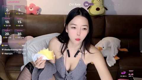 Media: Video of a young East Asian woman with long black hair, fair skin, and a slender physique, wearing a grey halter top and fishnet stockings, seated on a brown couch. Background features stuffed animals and a messy room.